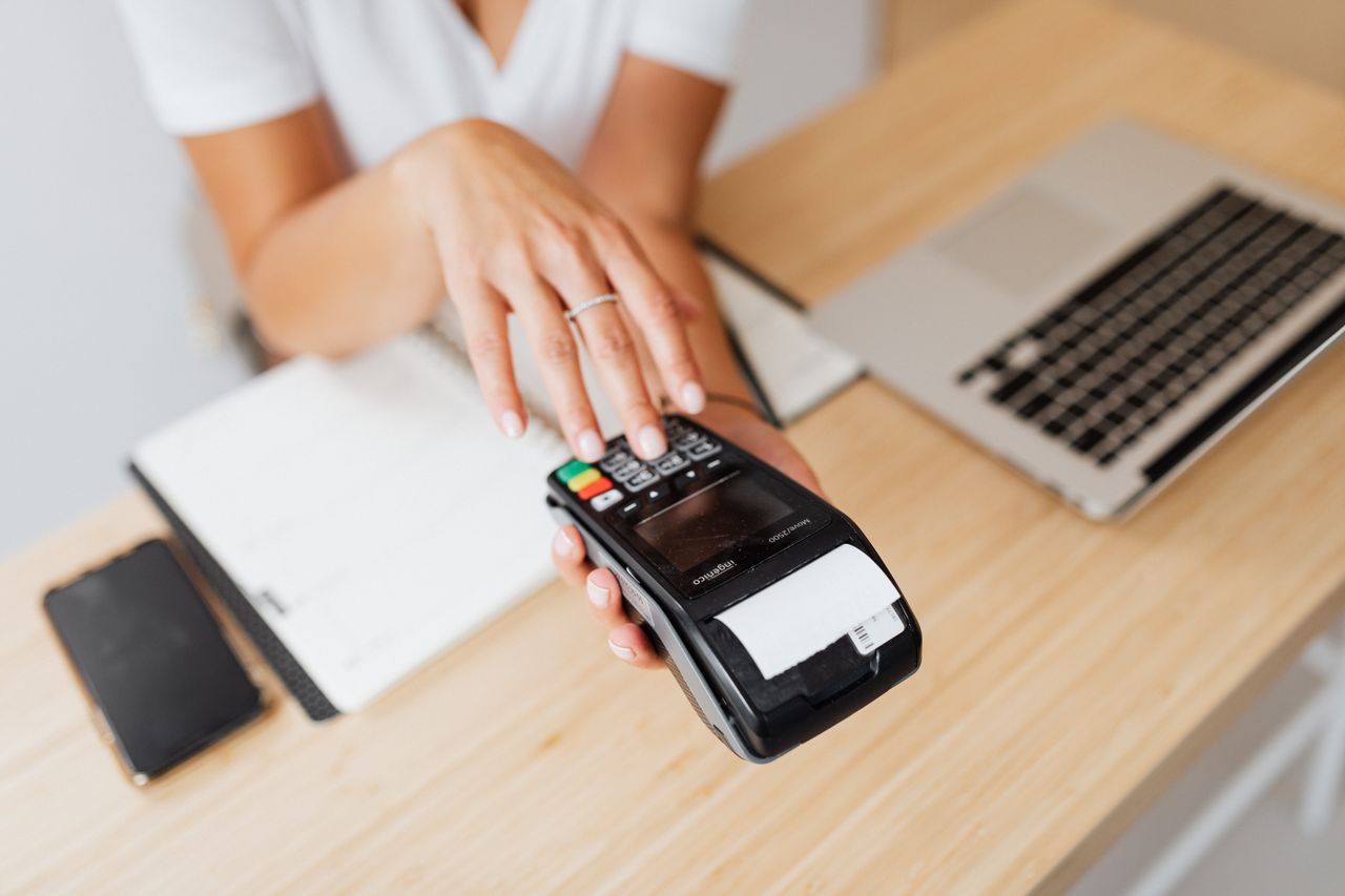 Home Close-up of a hand using a payment terminal beside a laptop on a wooden countertop.